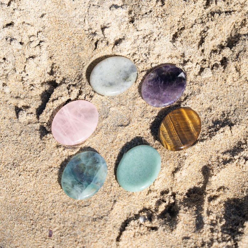 Puede incluir: Seis piedras preciosas pulidas de varios colores, incluyendo rosa, p&uacute;rpura y verde, dispuestas en c&iacute;rculo sobre arena. Las piedras tienen formas ovaladas lisas y patrones naturales. La arena proporciona un fondo neutro y texturizado.