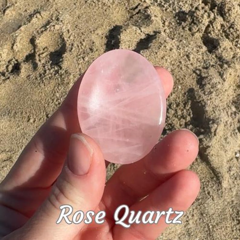Puede incluir: Una piedra de cuarzo rosa lisa y ovalada, sostenida en una mano. La piedra es de un color rosa p&aacute;lido con finas vetas internas. El texto "Rose Quartz" est&aacute; escrito en la parte inferior de la imagen.