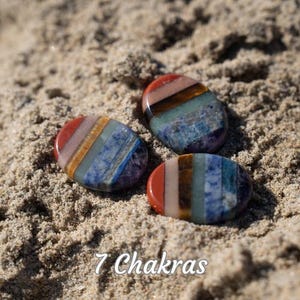Puede incluir: Tres piedras pulidas, de forma ovalada, con una gama de colores del arco&iacute;ris, incluyendo rojo, naranja, amarillo, verde, azul, &iacute;ndigo y violeta, se muestran sobre una superficie arenosa. El texto "7 Chakras" est&aacute; escrito en cursiva en la parte inferior de la imagen.