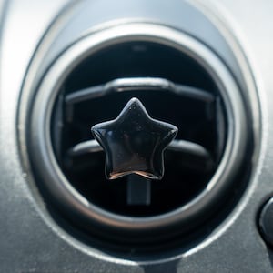 May include: A glossy black star-shaped air freshener clip attached to the horizontal slats of a circular car air vent. The star has a smooth, reflective surface and is centered within the dark gray plastic housing of the vehicle dashboard.
