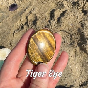 Puede incluir: Gema de ojo de tigre ovalada con rayas marr&oacute;n dorado y negras. La piedra se sostiene en una mano sobre un fondo arenoso. El texto "Tiger Eye" est&aacute; escrito en cursiva blanca en la parte inferior de la imagen.
