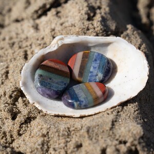 Puede incluir: Tres piedras de chakra ovaladas en una concha blanca sobre arena. Cada piedra presenta un espectro de colores, incluyendo rojo, naranja, amarillo, verde, azul, &iacute;ndigo y violeta. Las piedras son lisas y pulidas.