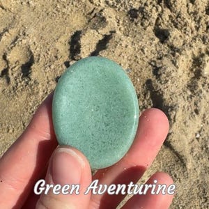 Puede incluir: Una piedra de aventurina verde lisa y de forma ovalada. La piedra es de color verde claro con una superficie ligeramente texturizada. El texto "Green Aventurine" est&aacute; escrito en blanco en la parte inferior de la imagen.