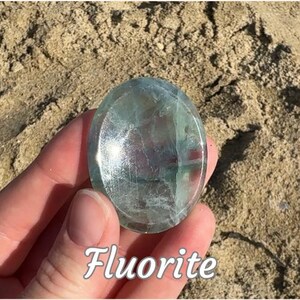 Puede incluir: Una piedra de fluorita pulida, de forma ovalada y aspecto transl&uacute;cido. La piedra presenta tonos verdes, azules y morados. La palabra "Fluorita" est&aacute; escrita en cursiva en la parte inferior de la imagen.