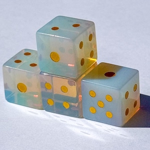 May include: Three translucent, opalescent dice with a milky, iridescent finish. Each cube features rounded corners and bright yellow circular pips. The dice are arranged in a small stack, showing various numerical faces with a smooth, polished surface.