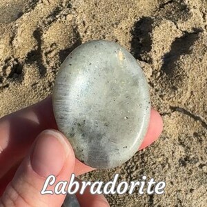 Puede incluir: Una piedra de labradorita pulida, de forma ovalada, de color gris p&aacute;lido con un brillo iridiscente. La piedra tiene motas de color gris oscuro y se sostiene sobre un fondo arenoso. La palabra "Labradorite" est&aacute; escrita en cursiva.