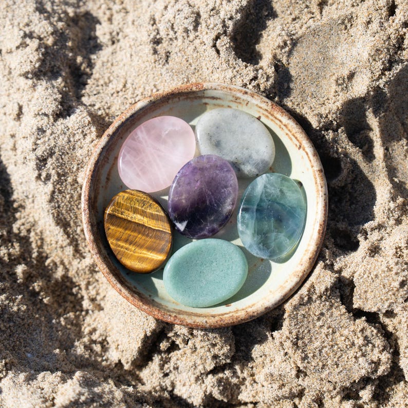 May include: A ceramic bowl filled with six polished gemstones of various colors, including pink, purple, green, and gold. The bowl sits on a sandy surface, with the gemstones arranged in a circular pattern. The gemstones are oval-shaped and appear smooth and reflective.