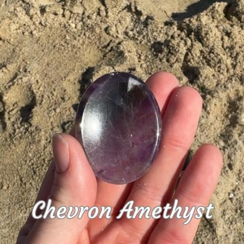 Puede incluir: Una piedra de amatista Chevron pulida, de forma ovalada, que muestra una mezcla de tonos morados y blancos. La piedra se sostiene en una mano sobre un fondo arenoso. El texto "Chevron Amethyst" est&aacute; escrito en blanco en la parte inferior de la imagen.