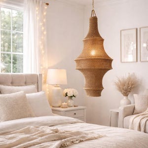 May include: A woven, tiered pendant light fixture hangs from a gold-colored ceiling mount. The light fixture is made of a natural fiber material and is suspended above a bed with neutral-toned bedding. The bedroom has a soft, inviting aesthetic.