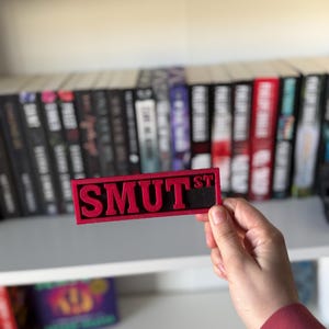 Op de afbeelding: Een rechthoekige patch met de tekst "SMUT ST" in rode letters op een zwarte achtergrond. De patch wordt voor een boekenplank vol boeken gehouden.