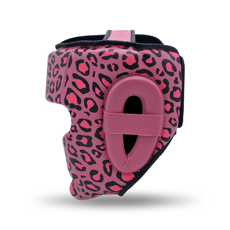 May include: A pink leopard print headgear with black accents. The headgear features a padded ear protector and a chin strap. The design includes a pink and black leopard print pattern, offering protection and style for combat sports.