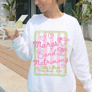 May include: White sweatshirt with a pink and green graphic that reads "Margs and Matrimony" with a ring and cocktail glass illustration. The text "Catie's Back, Palm Beach, 2026" is also included. The sweatshirt is a casual style.