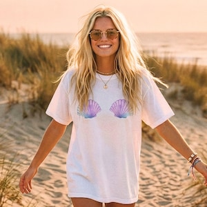May include: White t-shirt featuring a seashell design in shades of purple and blue. The shirt has a relaxed fit and short sleeves. The image shows a person wearing the shirt on a sandy beach.