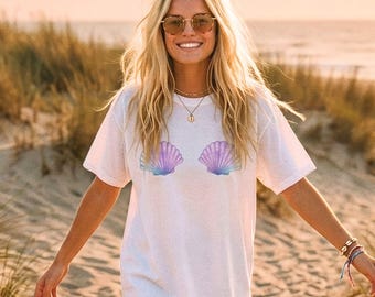 May include: White t-shirt with a seashell design in purple and blue. The shirt has a loose fit and short sleeves. The image shows a person wearing the t-shirt on a sandy beach.