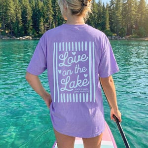May include: A light purple t-shirt with the words "Love on the Lake" in white script and a vertical striped design. The shirt is worn by a person on a paddleboard on a lake. The text "CATIE'S BACH LAKE TAHOE 20" is also visible.