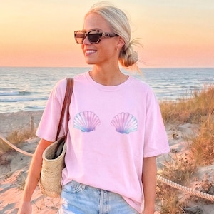 May include: Pink t-shirt featuring two seashell graphics in shades of pink, blue, and purple. The shirt is worn by a person on a beach, with the ocean and sunset in the background. The person is wearing sunglasses and carrying a straw bag.