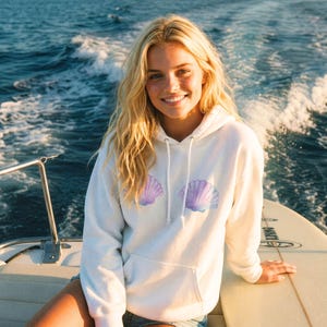 Puede incluir: Una sudadera con capucha blanca con un bolsillo delantero y un diseño de concha en tonos morados y azules. La sudadera se lleva en un barco con el océano de fondo.