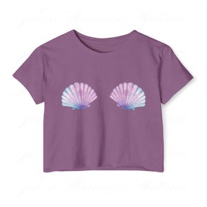 May include: A purple crop top with two seashell designs in blue, pink, and purple. The seashells are positioned centrally on the front. The top has short sleeves and a round neckline.