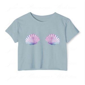 May include: A pale blue t-shirt with two seashell designs. The shells are in shades of pink, purple, and blue. The t-shirt has short sleeves and a round neck. The design is positioned on the chest.
