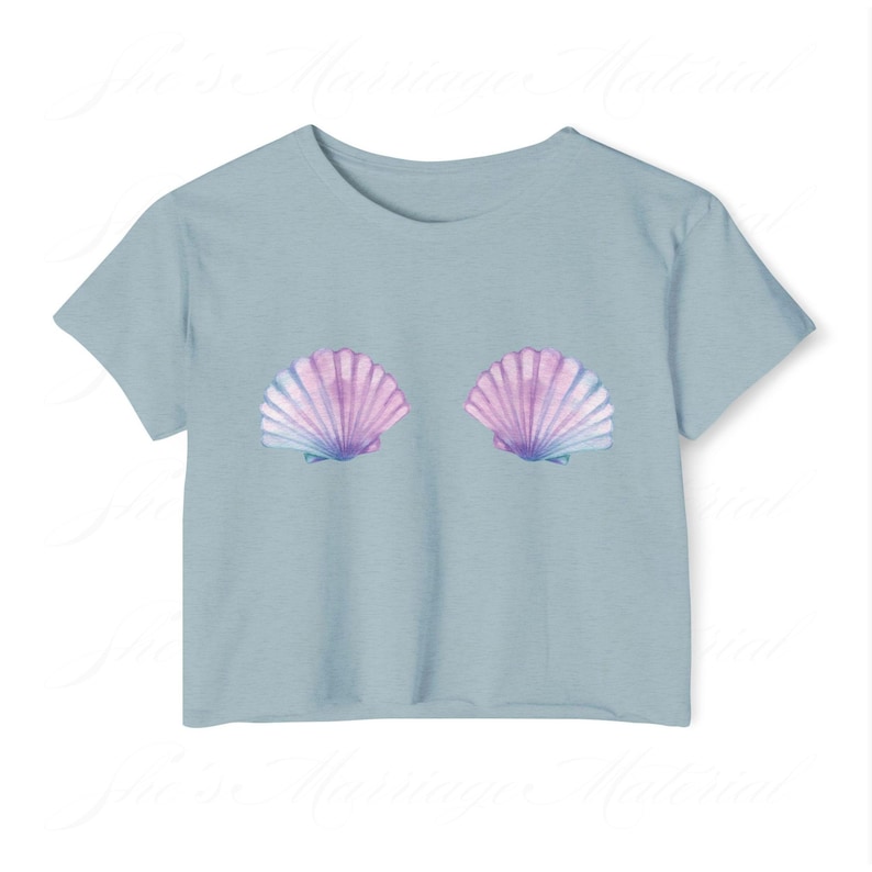 May include: A pale blue t-shirt with two seashell designs. The shells are in shades of pink, purple, and blue. The t-shirt has short sleeves and a round neck. The design is positioned on the chest.