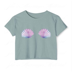 May include: A pale blue t-shirt with two seashell illustrations in purple, pink, and blue. The shells are positioned centrally on the front. The top has short sleeves and a round neckline, offering a casual and summery look. The design is reminiscent of the sea.