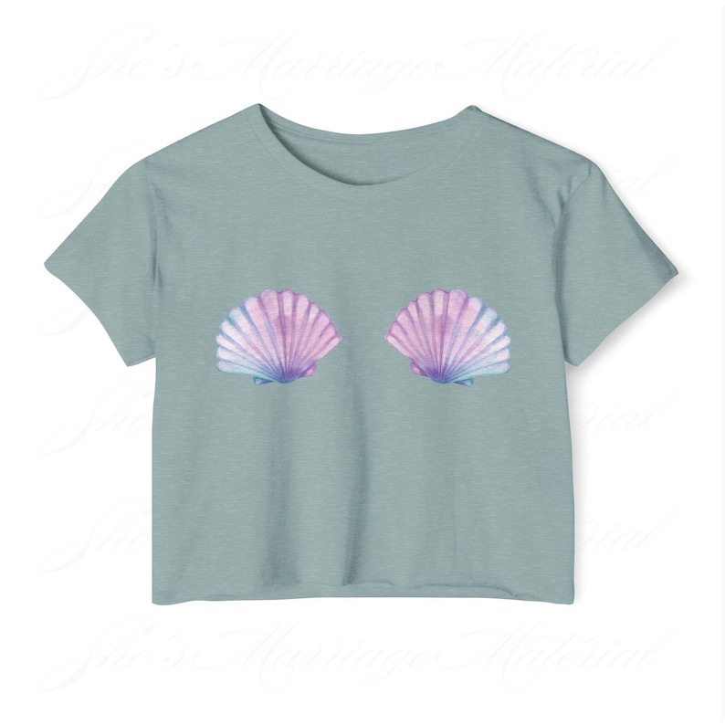 May include: A pale blue t-shirt with two seashell illustrations in purple, pink, and blue. The shells are positioned centrally on the front. The top has short sleeves and a round neckline, offering a casual and summery look. The design is reminiscent of the sea.
