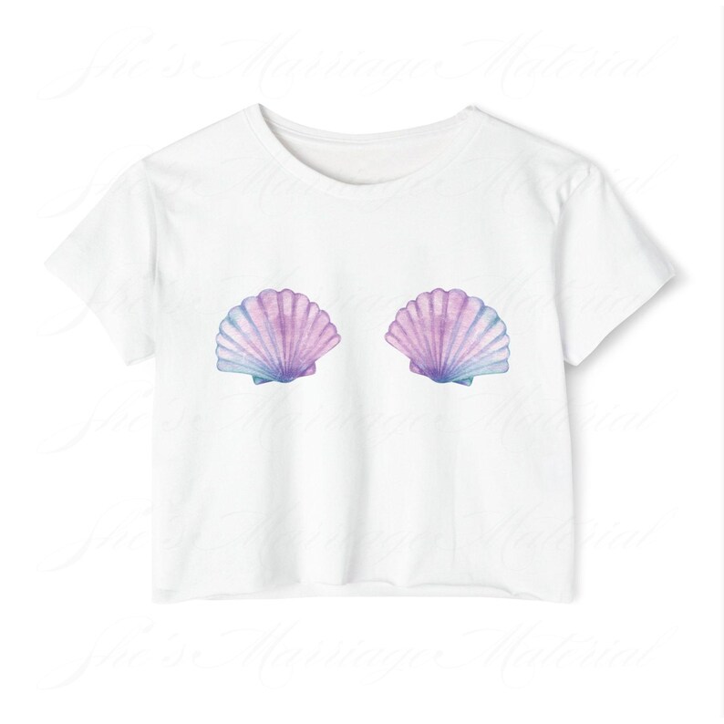 May include: White t-shirt with a watercolour design of two seashells. The shells are in shades of purple, blue, and pink. The shirt has a classic crew neck and short sleeves. The design is centered on the chest.