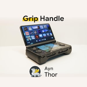 May include: A black handheld gaming console with a dual-screen clamshell design, featuring a top screen displaying a grid of game icons and a bottom screen showing gameplay. The text Grip Handle and Ayn Thor are visible along with a dragon logo.
