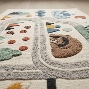 Puede incluir: Una alfombra de color crema con un dise&ntilde;o de camino sinuoso en gris texturizado. La alfombra incluye varios motivos en relieve como una cara de oso marr&oacute;n, mariquitas rojas, formas geom&eacute;tricas azules, una ballena y elementos de paisaje verdes.