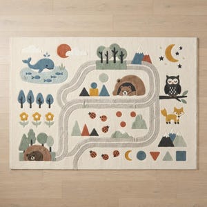 Puede incluir: Una alfombra rectangular con fondo crema que presenta un patr&oacute;n de carretera gris sinuosa. El dise&ntilde;o incluye una ballena azul, un oso pardo en una cueva, un oso durmiendo, un b&uacute;ho, un zorro, mariquitas, &aacute;rboles, monta&ntilde;as y formas geom&eacute;tricas.