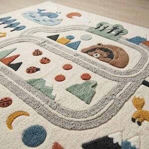 Puede incluir: Una alfombra de color blanco roto con un dise&ntilde;o texturizado y acolchado de tem&aacute;tica natural. El patr&oacute;n incluye un oso pardo durmiendo en una cueva, una ballena azul, mariquitas, un zorro, monta&ntilde;as, &aacute;rboles, una luna creciente y un camino gris.