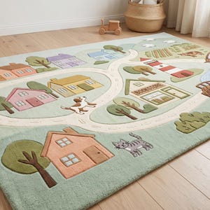 Puede incluir: Una alfombra rectangular con fondo verde claro que presenta una escena urbana. El diseño incluye casas de varios colores, un camino sinuoso, un faro, una librería, árboles, un perro, un gato y un río azul con un puente de madera.