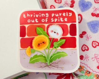 May include: A square sticker featuring a cartoon illustration of dandelions growing in front of a red brick wall. The sticker has the text "thriving purely out of spite" in white letters. The dandelions are yellow and white with green leaves.
