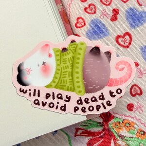 May include: A pink sticker featuring a cartoon rat with a white head, green sweater, and purple body. The sticker reads "will play dead to avoid people" in black cursive font.