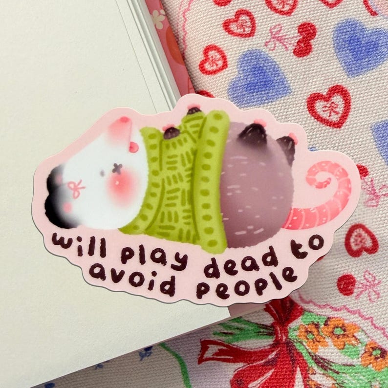 May include: A pink sticker featuring a cartoon rat with a white head, green sweater, and purple body. The sticker reads "will play dead to avoid people" in black cursive font.