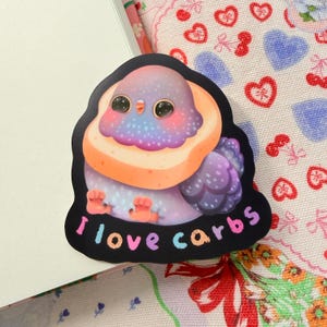 May include: A colorful sticker featuring a cartoon pigeon with a bread-like halo. The pigeon is primarily purple, with orange and pink accents. The text "I love carbs" is written in a rainbow font at the bottom of the sticker.