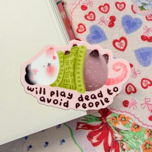 May include: A pink sticker with a cartoon illustration of a creature playing dead. The creature is white, green, and purple, with a pink tail. The sticker has the text "will play dead to avoid people."