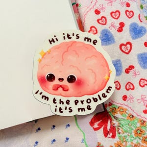May include: A sticker featuring a cartoon brain with a cute, blushing face and the text "Hi it's me, I'm the problem, it's me." The brain is pink with yellow star accents.