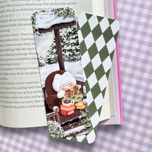 May include: Two rectangular bookmarks. One features a white cat wearing glasses, reading a book in a brown armchair on a snowy porch with stacked books and a mug. The other bookmark has a repeating dark green and white diamond argyle pattern.