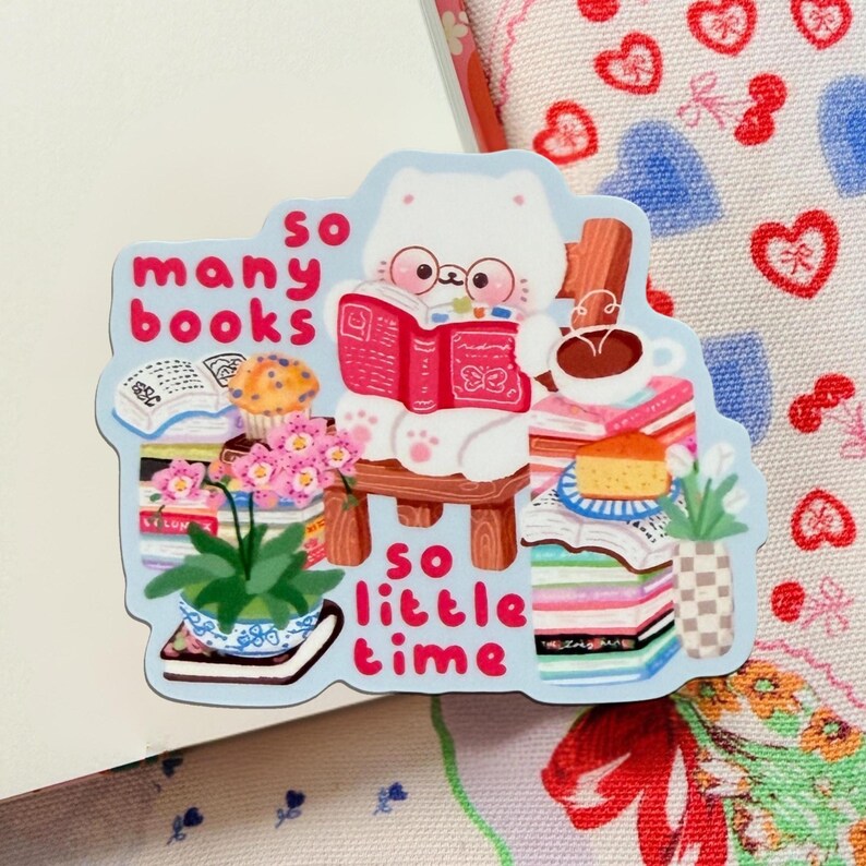 May include: A sticker depicting a white cat wearing spectacles, reading a red book, and sitting on a wooden chair. The sticker includes the text "so many books so little time" and is surrounded by books, a cupcake, and a coffee cup.