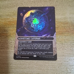 May include: A collectible card featuring a cosmic illustration of a planet with blue and green hues, titled "Gemstone Caverns." The card has text describing its gameplay mechanics and is a "Legendary Land" card.