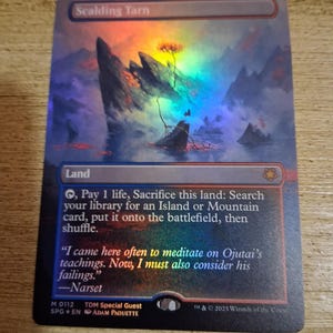 May include: A holographic Scalding Tarn card from the Magic: The Gathering trading card game. The card features a landscape with mountains, a tree, and text. The card's border is red and the text includes the card's name, rules, and flavor text.
