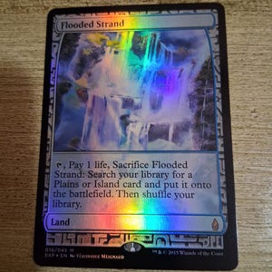 May include: A holographic "Flooded Strand" card from Magic: The Gathering. The card features a waterfall scene with a rainbow sheen. Text includes card name, instructions, and copyright information. The card is a Land card.