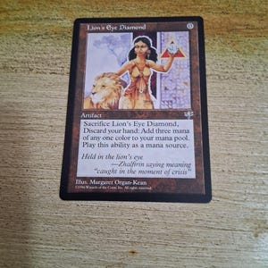 Puede incluir: Una carta de Magic: The Gathering titulada "Lion's Eye Diamond". La carta presenta una ilustración de una mujer con un león, sosteniendo un diamante. El texto de la carta describe las habilidades del artefacto e incluye el nombre del artista y la información de derechos de autor.