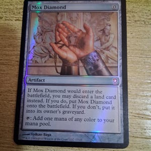 May include: A collectible Mox Diamond card with a holographic finish. The card features an illustration of hands holding a pendant, with text including "Artifact" and instructions for gameplay. The card's border is black.