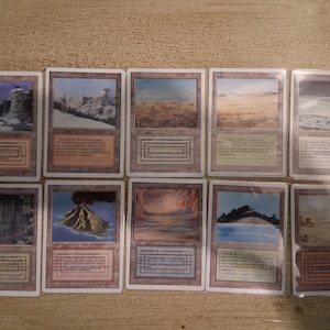 Op de afbeelding: Een verzameling van tien Magic: The Gathering ruilkaarten. De kaarten tonen verschillende landschapsillustraties, waaronder bergen, woestijnen en vulkanische eilanden. Elke kaart heeft een unieke rand en tekstvak.