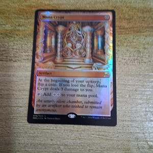 Puede incluir: Una carta holográfica de Magic: The Gathering titulada "Mana Crypt". La carta presenta una ilustración de una cámara detallada con pilares y arquitectura ornamentada. El texto describe su tipo de artefacto y sus efectos.