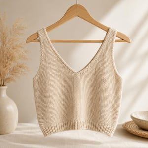 Peut inclure: Un débardeur en tricot beige sans manches avec une encolure en V profonde et un ourlet côtelé. Le vêtement est réalisé avec un point de jersey simple et est présenté suspendu à un cintre en bois, mettant en valeur ses lignes épurées.