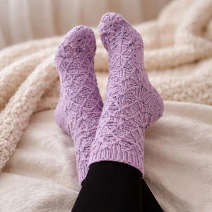 Puede incluir: Un par de calcetines de punto color lavanda con un patrón de diamantes. Los calcetines se llevan puestos, con pantalones negros visibles debajo. El fondo presenta una manta color crema.
