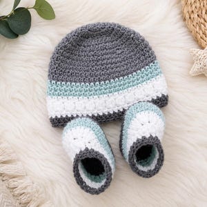 Peut inclure: Ensemble fait main comprenant un bonnet et des chaussons pour bébé au crochet. Le bonnet et les chaussons sont gris, blancs et bleu clair. L'ensemble est en fil doux et idéal pour un nouveau-né. Les chaussons mesurent environ 10 cm de long.